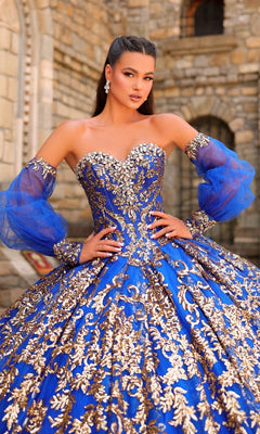 A woman poses in the Sequin Quinceanera Dress: Amarra 54238, a blue gown with gold embroidery, sheer puff sleeves, and a corset bodice, standing before a stone building.