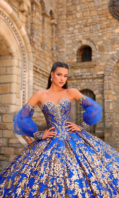 A woman poses by a stone building in the Sequin Quinceanera Dress: Amarra 54238, featuring a blue and gold corset bodice, puff sleeves, sheer accents, and ornate embroidery.