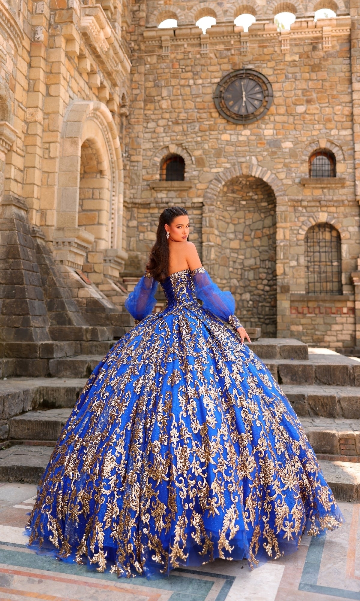 A woman in the Sequin Quinceanera Dress: Amarra 54238, a blue and gold ornate ball gown with puff sleeves, stands on stone steps before a large stone building with arched windows and a clock.