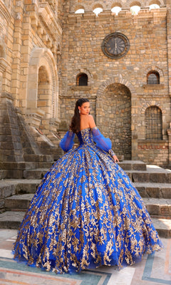 A woman in the Sequin Quinceanera Dress: Amarra 54238, a blue and gold ornate ball gown with puff sleeves, stands on stone steps before a large stone building with arched windows and a clock.