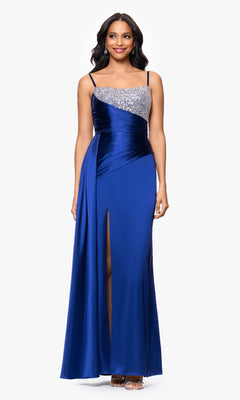 A woman wearing the Blondie Nites 5423BN long blue formal dress with a beaded bodice, thin straps, side drape, and front slit stands against a plain white background.