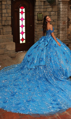 A woman poses indoors in the Amarra 54243 Off-Shoulder Quinceanera Dress, featuring an elaborate blue ball gown with 3D floral appliques, silver accents, and a long train, set against a wooden door and stone walls.