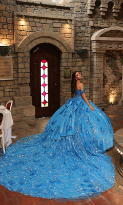 A woman wearing the Off-Shoulder Quinceanera Dress: Amarra 54243, a bright blue ball gown with 3D floral appliques and a long train, smiles over her shoulder in front of a wooden door in a stone-walled room.