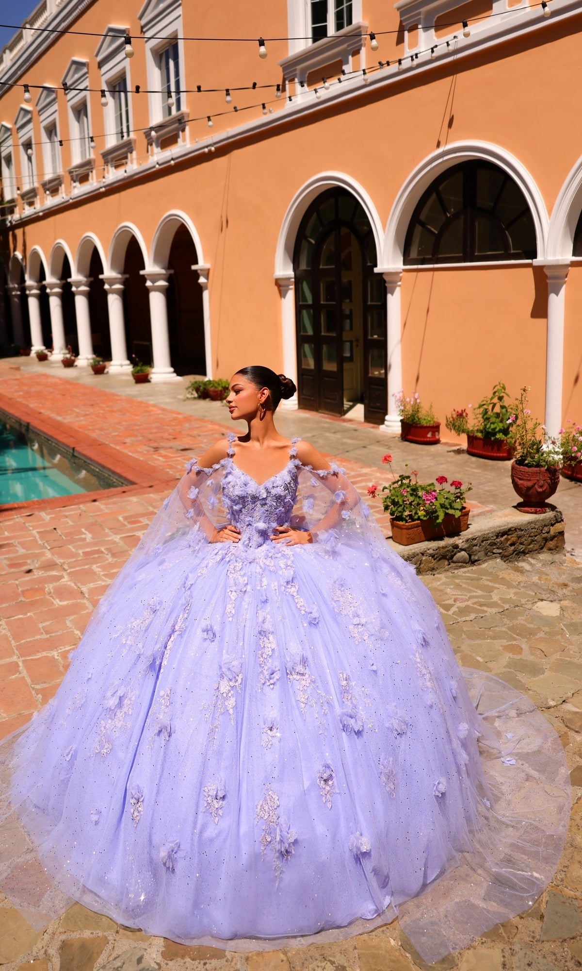 A person stands by a pool in a courtyard, wearing the Amarra 54254 Periwinkle Purple Quincea¤era Dress with floral details and sheer sleeves.