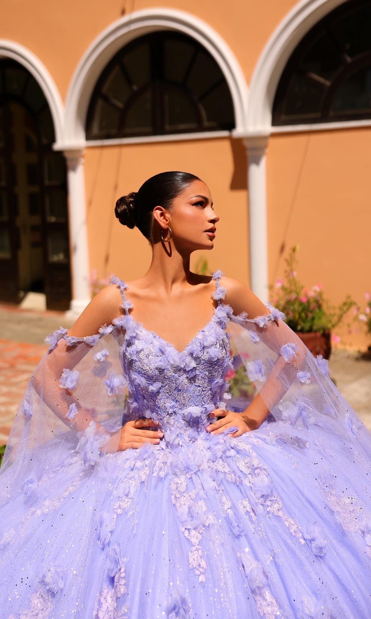A woman in the Amarra 54254 Periwinkle Purple Quincea¤era Dress with floral details and sheer sleeves stands outside an arched building, gazing to the side.
