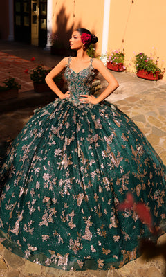 A young woman poses outdoors wearing the Amarra 54260 Corset Quincea¤era Dress with Cloak?a green ball gown with silver floral designs?hands on hips, in front of a peach-colored wall and potted plants.
