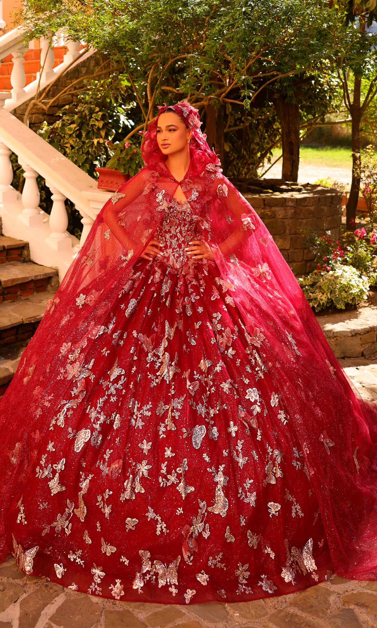 A woman poses confidently outdoors in the Amarra 54260 Corset Quincea¤era Dress with Cloak?a voluminous red ball gown featuring silver floral patterns and a matching cape.
