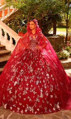 A woman poses confidently outdoors in the Amarra 54260 Corset Quincea¤era Dress with Cloak?a voluminous red ball gown featuring silver floral patterns and a matching cape.