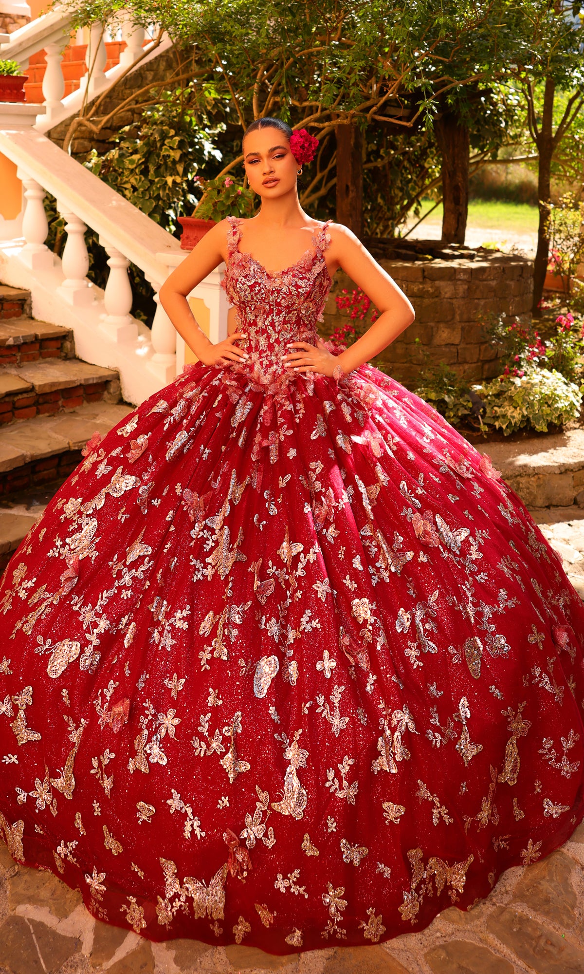 A woman stands outdoors in front of stairs, wearing the Amarra 54260 Corset Quincea¤era Dress with Cloak?a voluminous red ball gown featuring silver floral and butterfly patterns.