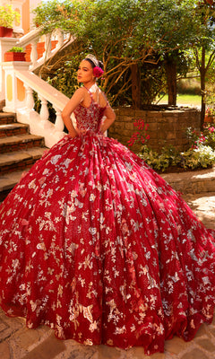 A young woman poses outdoors on a stone path, wearing the Amarra 54260 Corset Quincea¤era Dress with Cloak?a voluminous red gown with silver floral patterns and a tiara?near stairs and greenery.