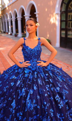 A young woman poses outside in the Amarra 54260 Corset Quincea¤era Dress with Cloak, a formal blue gown adorned with floral appliqu‚s and glitter, her hair up with a white flower accessory and hands on hips.
