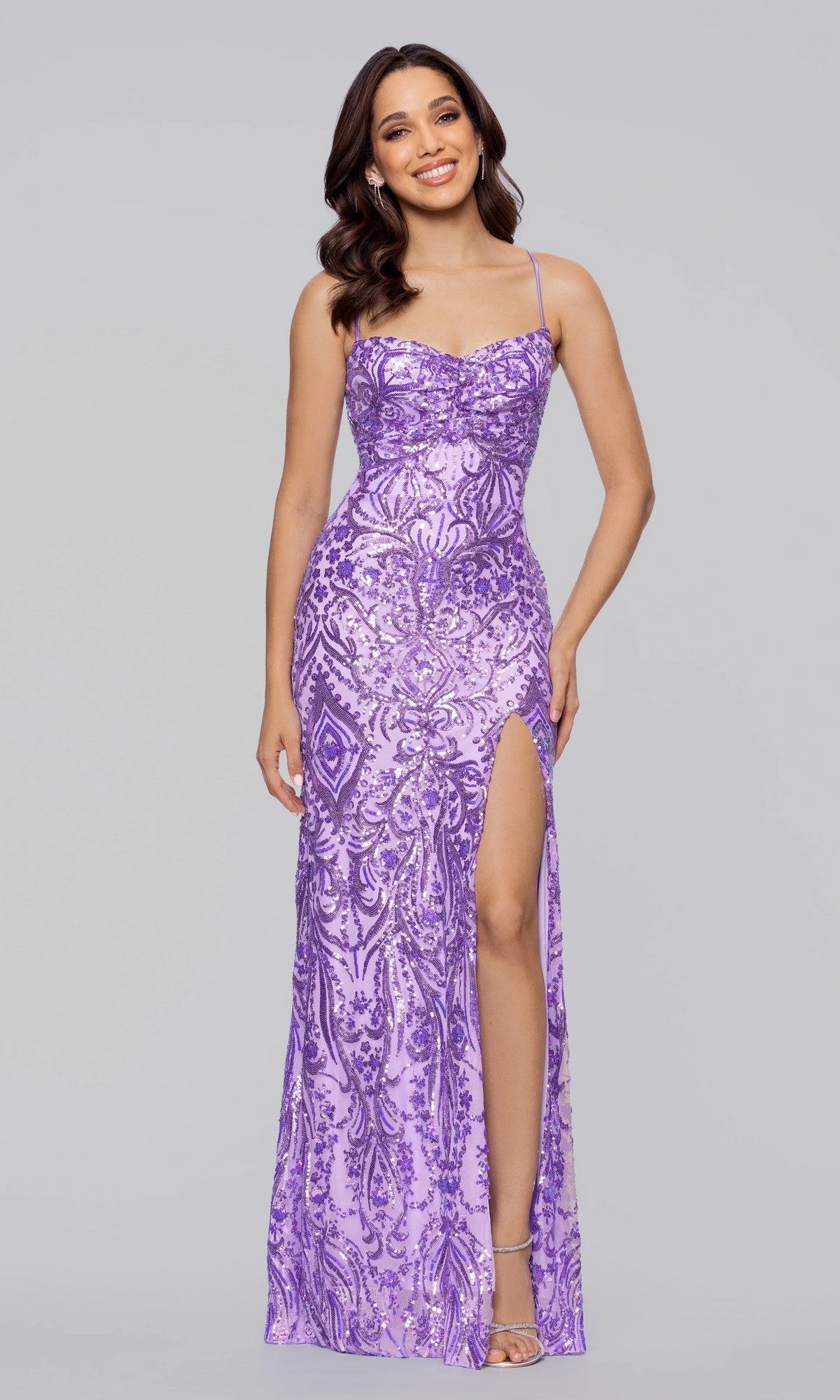 A woman smiles at the camera in the Blondie Nites 5428BN long prom dress, featuring lavender sequins, spaghetti straps, and a high leg slit, standing against a plain gray background.