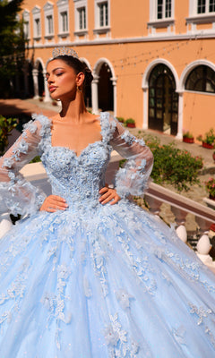 Woman wearing the Corset Ball Gown Quinceañera Dress 59290 by Amarra, a blue glitter tulle gown with floral embroidery and tiara, poses hands on hips before an elegant peach building with arched windows.