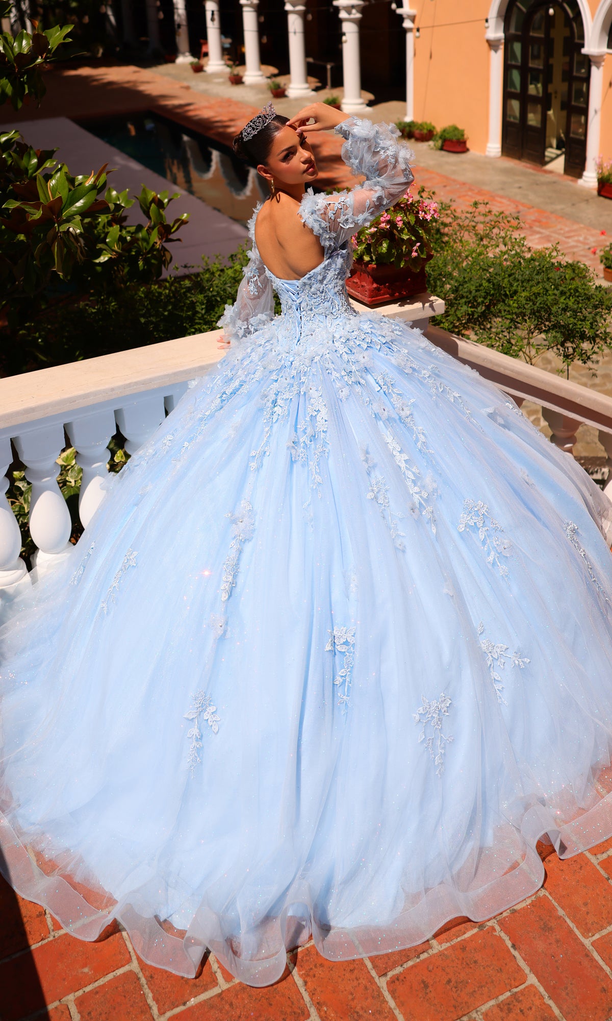 A young woman in the Corset Ball Gown Quinceañera Dress 59290 by Amarra, featuring glitter tulle and a tiara, poses on a balcony with white railings and greenery in the background.