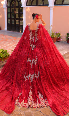 A woman stands outdoors, seen from the back in the Amarra 54296 Red and Gold Quincea¤era Dress with silver embroidery. She holds one hand to her head, flowers in her hair, as the elegant gown cascades gracefully behind her.