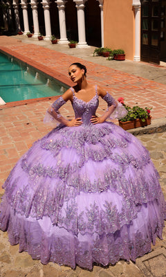A woman stands by a pool wearing the Quinceanera Dress 54300 By Amarra, a voluminous lavender ball gown with glittery floral accents and removable sheer sleeves.