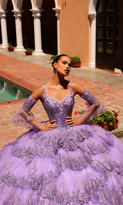 A woman poses by a pool in the Quinceanera Dress 54300 By Amarra, a detailed sparkling lavender ball gown with lace embellished removable sheer sleeves, surrounded by brick pavement, potted plants, and arches.