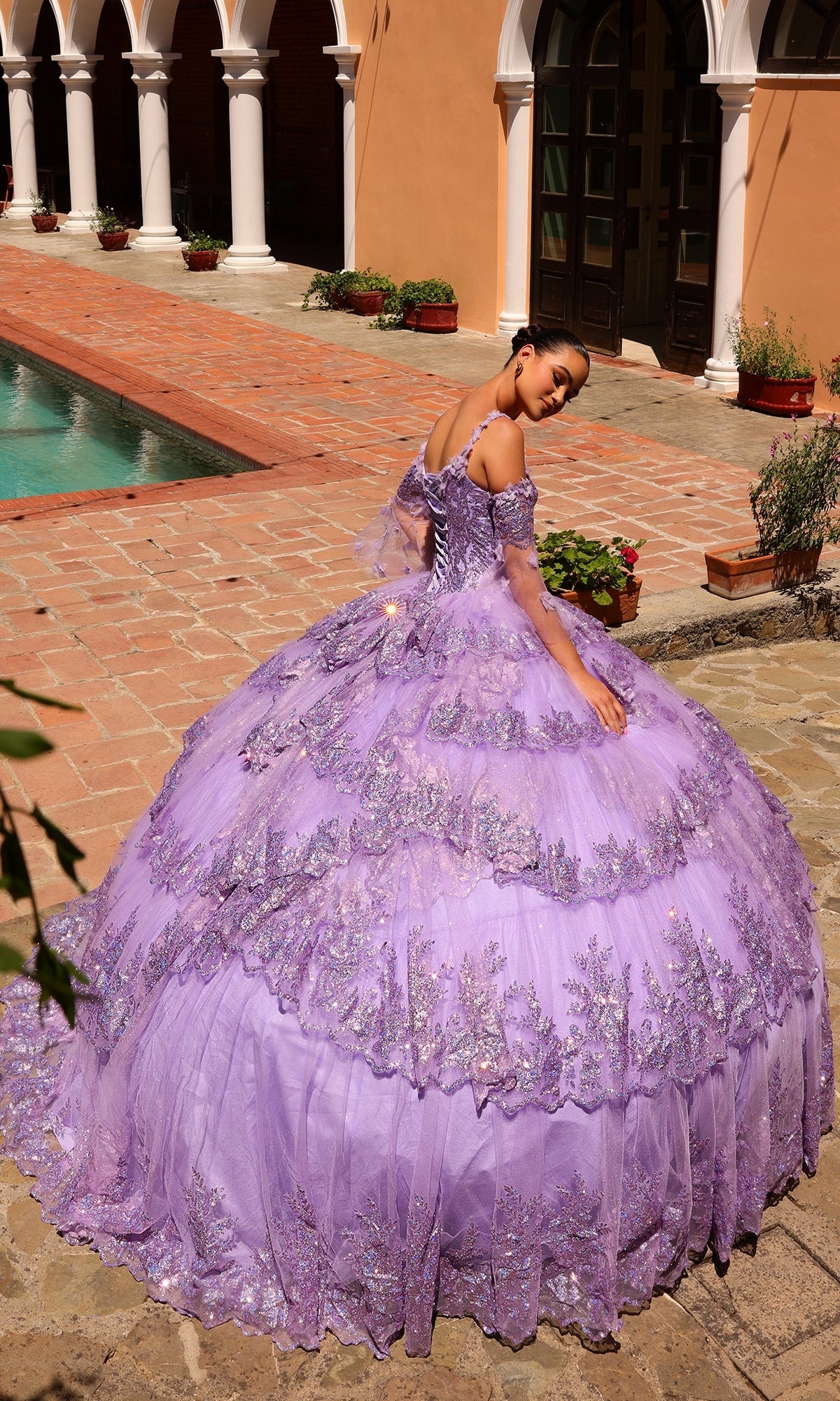 A young woman in the Quinceanera Dress 54300 By Amarra, a lace embellished lavender ball gown with removable sheer sleeves, stands near a pool and an arched building.
