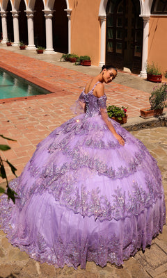 A young woman in the Quinceanera Dress 54300 By Amarra, a lace embellished lavender ball gown with removable sheer sleeves, stands near a pool and an arched building.