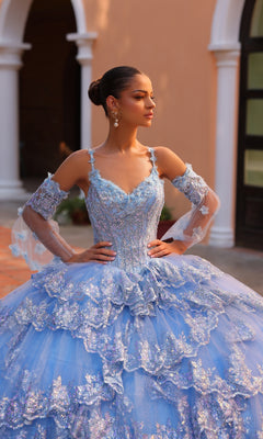 Wearing the Quinceanera Dress 54300 By Amarra, a young woman stands outdoors in a sparkly blue ball gown with lace details and removable sheer sleeves, hands on her hips as she gazes to the side.