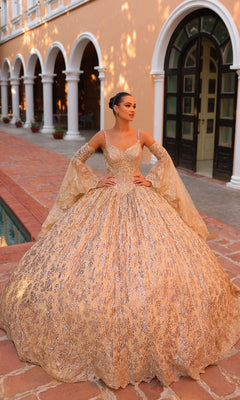 A woman stands outside a building wearing the Quinceanera Dress 54303 By Amarra: an ornate, gold, floor-length v-neck ball gown with flared sleeves.