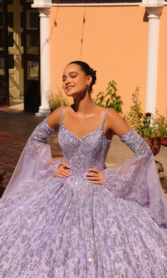 A woman poses outdoors in the sunlight wearing the Quinceanera Dress 54303 By Amarra, a sparkling lavender sequin ball gown with off-the-shoulder sleeves, standing before a peach building with white columns.