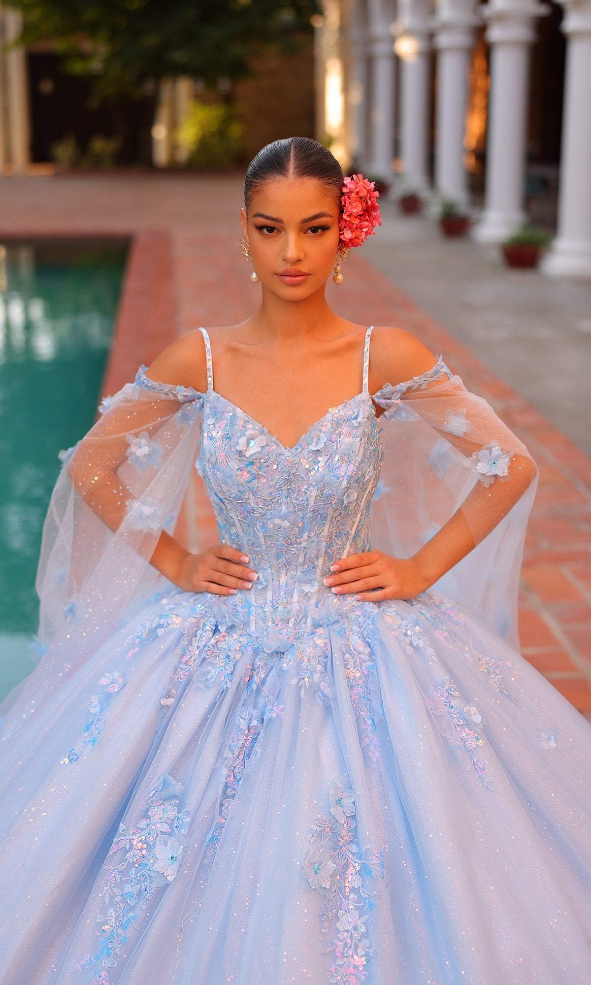 Wearing the Quinceanera Dress 54306 By Amarra, a woman with a pink flower in her hair and hands on her hips stands by a pool in a light blue ball gown featuring a corset bodice and sheer sleeves.