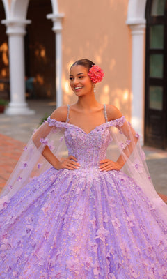 A young woman smiles outside in the Quinceanera Dress 54308 By Amarra, an off-the-shoulder ball gown with 3D floral details, a sheer cape, and a large pink flower in her hair.