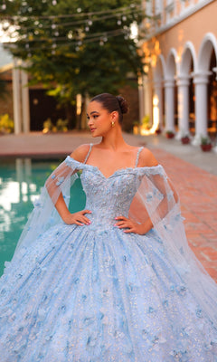 A young woman stands by a pool, wearing the Amarra Quinceanera Dress 54308, an off-the-shoulder light blue ball gown featuring sheer cape sleeves and elegant 3D floral details.