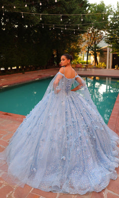 A woman in the Quinceanera Dress 54308 By Amarra?a blue, glittering off-the-shoulder ball gown with sheer cape sleeves?stands by a pool on a brick patio, facing away under string lights.