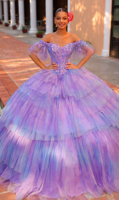 A young woman stands outdoors wearing the Quinceanera Dress 54312 by Amarra, a sparkly off-the-shoulder ball gown featuring layered tulle, a sweetheart neckline, and a pink flower in her hair.