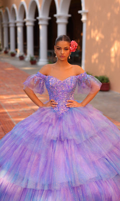 A young woman wearing the Quinceanera Dress 54312 By Amarra, a sparkling purple ball gown with a sweetheart neckline and off-the-shoulder sleeves, stands outdoors on a brick pathway near an arched building, with a pink flower in her hair.
