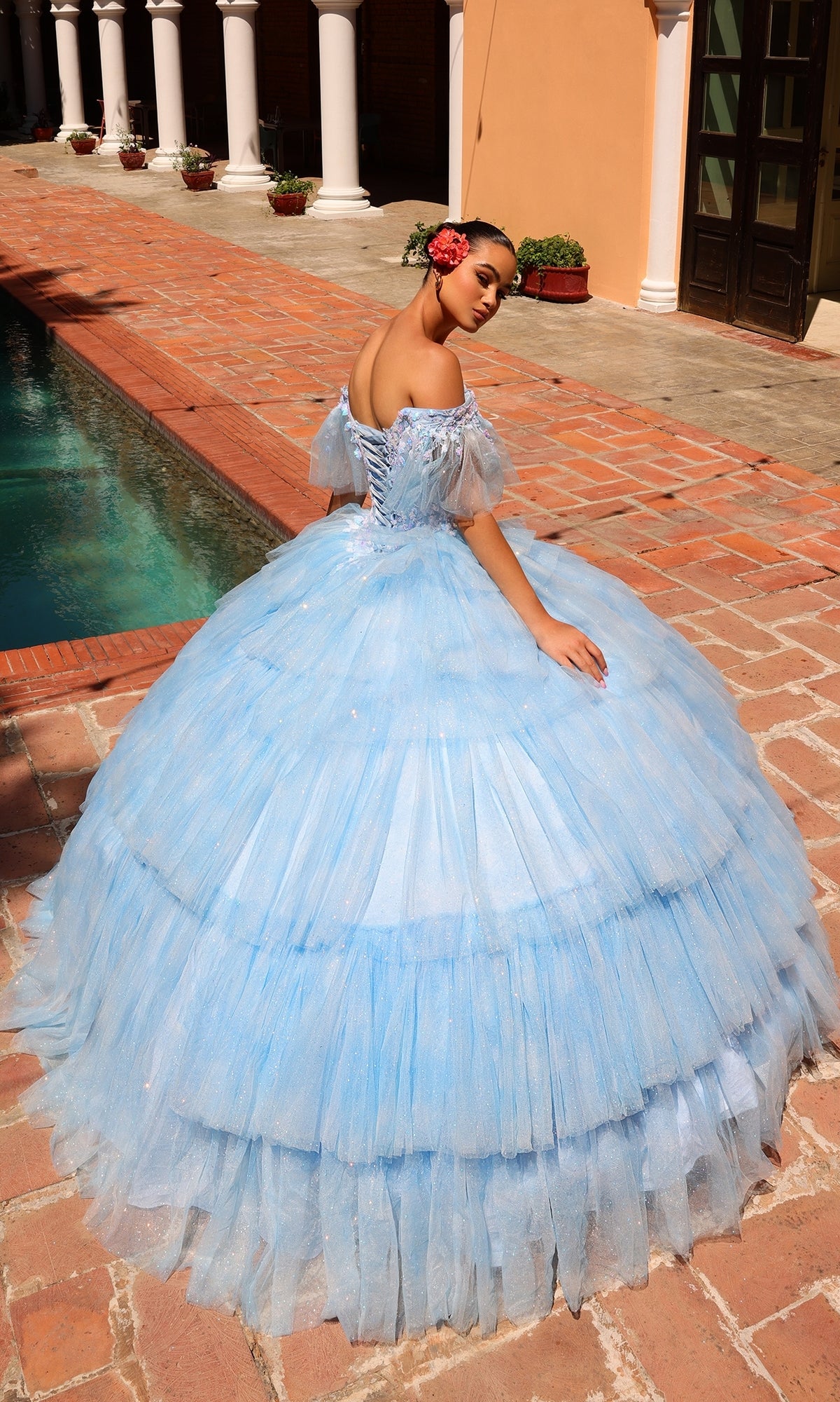 A woman in the Quinceanera Dress 54312 By Amarra, a light blue off-the-shoulder ball gown with a sweetheart neckline, stands on a brick walkway beside a pool, glancing back over her shoulder.