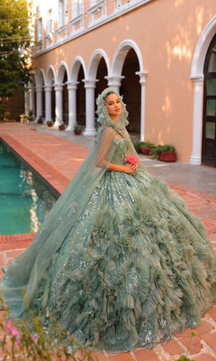 By the pool, someone poses in the Quinceanera Dress 54314 By Amarra, an elaborate sage green ball gown with floral details and a matching sheer cape train, holding a pink bouquet.