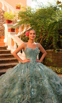A young woman poses outdoors in the Quinceanera Dress 54314 By Amarra, a light blue-green ball gown featuring floral accents, a sheer cape train, and a pink flower in her hair, with her hands on her hips.