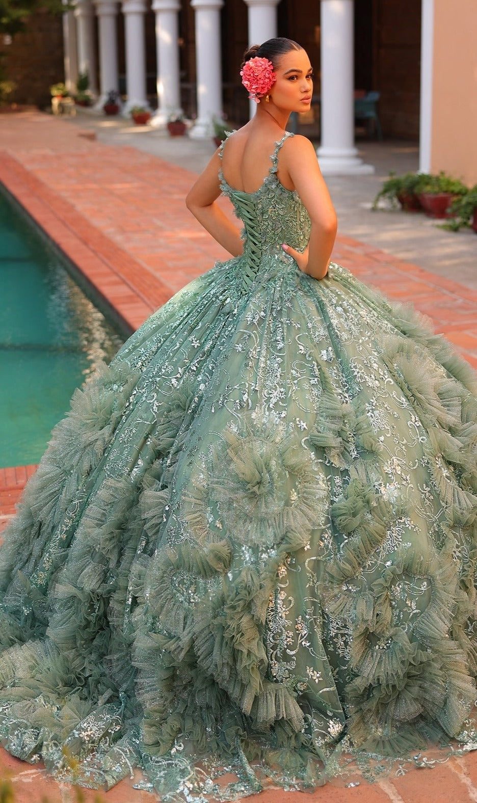 By the pool, a woman poses in the Quinceanera Dress 54314 by Amarra?a green v-neck ball gown featuring lace and ruffle details with a sheer cape train. Her bun is adorned with a large pink flower.