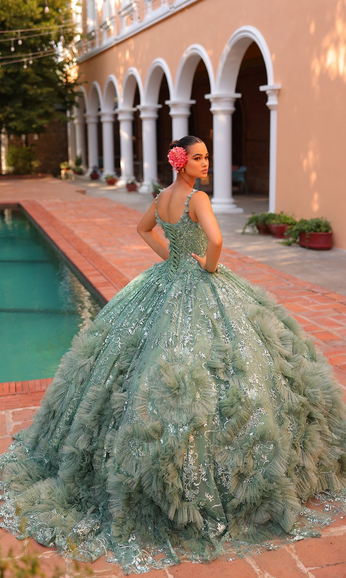 A young woman stands near a pool, facing away, in the Quinceanera Dress 54314 By Amarra?a large green ornate ball gown with sheer cape train. She wears a pink flower in her hair outside an arched building.