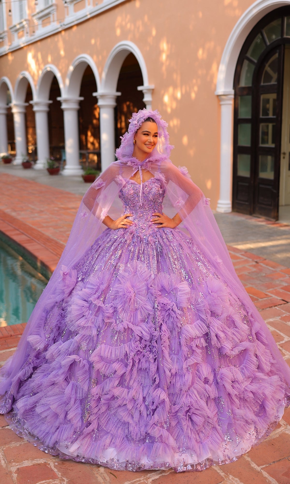 A woman poses outdoors in front of an arched building wearing the Amarra Quinceanera Dress 54314?a purple ball gown with layered ruffles, sparkles, a sheer cape train, and matching headpiece.