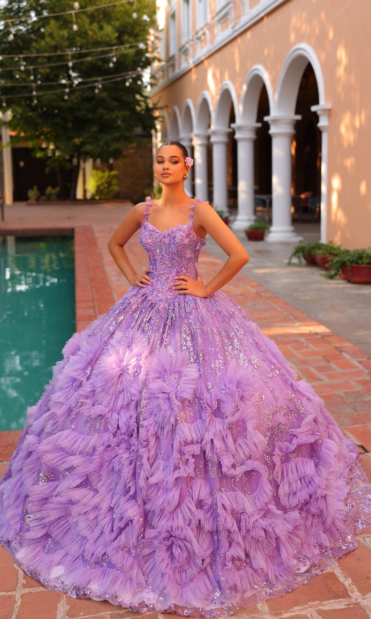Wearing the Quinceanera Dress 54314 by Amarra, a young woman poses by a pool in an elaborate lavender v-neck ball gown with floral details and a sheer cape train, her hands resting on her hips.