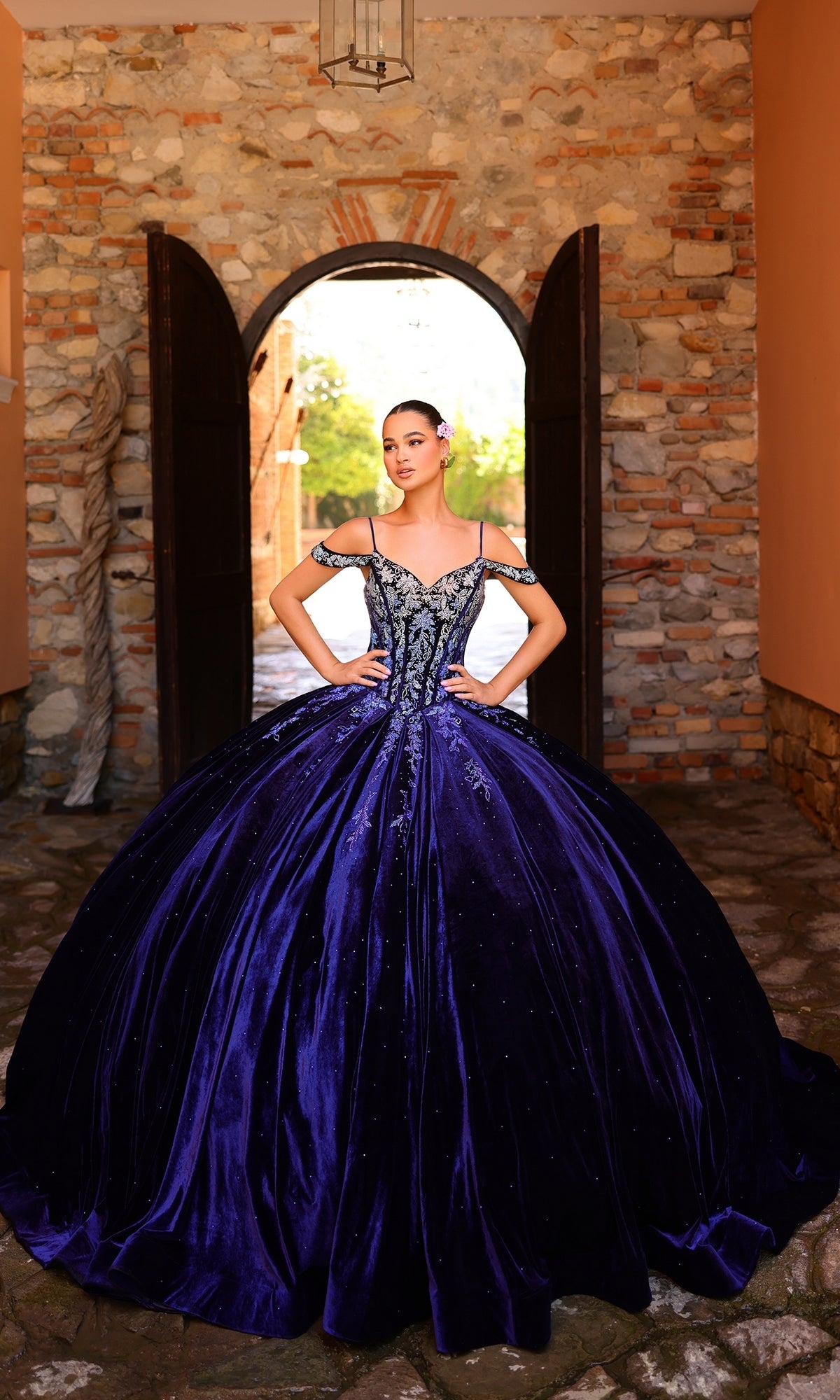 A woman stands before an arched doorway and stone walls, wearing the Quinceanera Dress 54316 By Amarra?a navy blue off-the-shoulder ball gown with a full skirt, ornate bodice, and ombre lace embroidery.