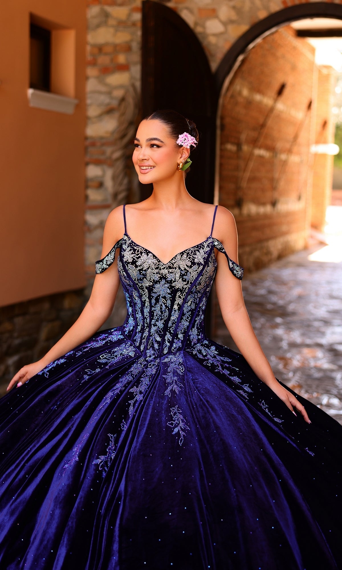 A young woman wears the Quinceanera Dress 54316 by Amarra?an off-the-shoulder ball gown with ombre lace embroidery?smiling outdoors with a pink flower in her hair and a stone archway behind her.