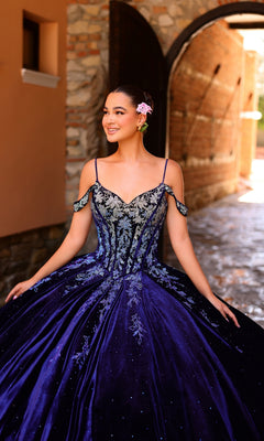 A young woman wears the Quinceanera Dress 54316 by Amarra?an off-the-shoulder ball gown with ombre lace embroidery?smiling outdoors with a pink flower in her hair and a stone archway behind her.