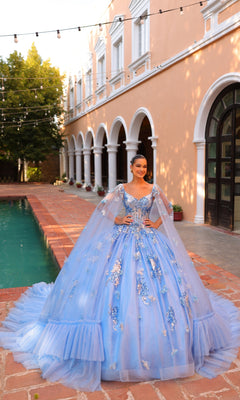 A young woman wearing the Quinceanera Dress 54318 By Amarra, a blue ball gown with floral embroidery, stands by a pool near a peach-colored building with arches and string lights overhead.