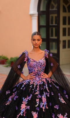 A woman stands outdoors in front of a building, wearing the Quinceanera Dress 54318 by Amarra?a black ball gown with pink and purple floral embroidery and sheer black sleeves?her hands resting on her waist.