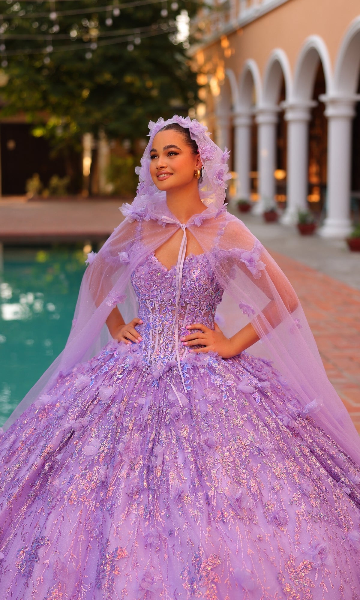 A young woman stands by a pool wearing the Quinceanera Dress 54320 by Amarra, an off-the-shoulder lavender ball gown with a matching cape and hood, featuring beautiful 3D floral details.
