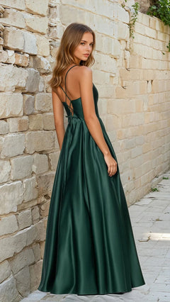 A woman in the Classic Long Prom Dress: Blondie Nites 5446BN, a dark green satin floor-length a-line gown with spaghetti straps and a lace-up open back, stands on a stone walkway by a rough stone wall, glancing over her shoulder.