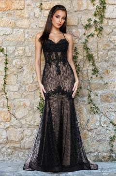 A woman wears the Blondie Nites 5463BN Long Black Lace Prom Dress in Black/Nude, standing before a stone wall covered with climbing green vines.