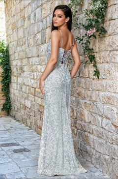 Wearing the Blondie Nites 5472BN long green sequin prom dress, a woman stands outdoors on a stone pathway beside a vine-covered wall, looking over her shoulder.
