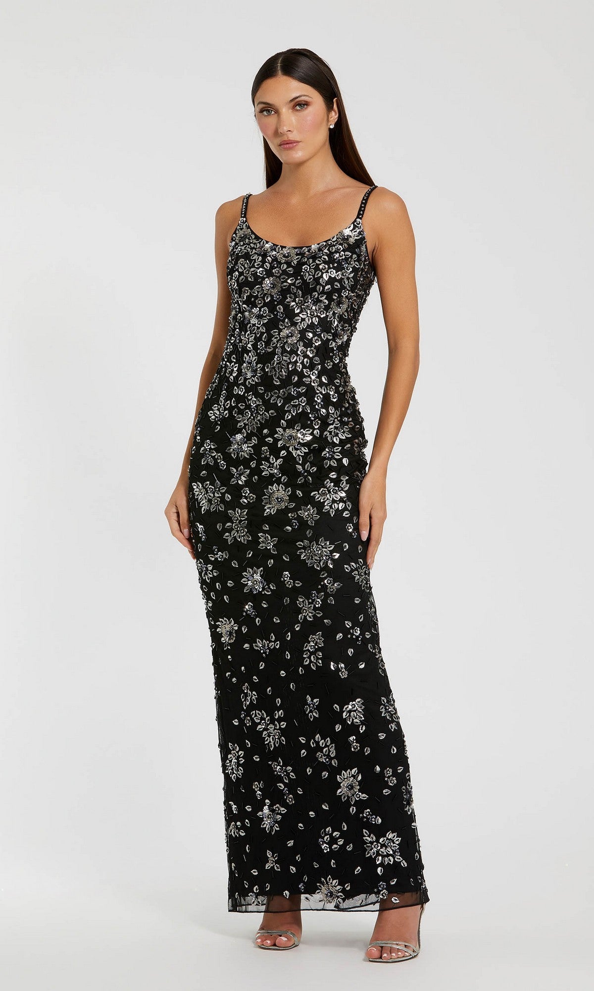 A woman in the Mac Duggal 5477 Floral-Beaded Long Formal Dress, featuring sleeveless design and silver embellishments, stands against a plain light background.
