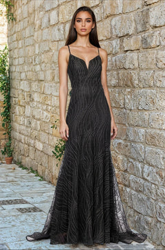 A woman stands outdoors against a stone wall, wearing the Long Black Glitter Prom Gown (Blondie Nites 5491BN), a fitted formal dress with thin straps and a subtle wavy pattern in black.